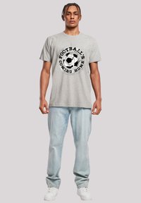 Gray cotton t-shirt featuring a black printed soccer ball graphic and text "FOOTBALL'S COMING HOME," paired with light blue jeans and white sneakers.