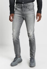 Jeans de mezclilla gris con un ajuste slim, que presentan detalles desgastados, un desvanecimiento sutil y un diseño clásico de cinco bolsillos. Llevados con zapatillas grises.