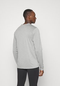 Ανοιχτό γκρι μακρυμάνικο πουκάμισο με στρογγυλεμένη λαιμόκοψη, εφαρμοστή σχεδίαση, απαλό ύφασμα και ελαφρώς στενότερα μανίκια, φορεμένο με μαύρο κολάν.