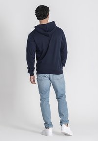 Navy modrý mikina s kapucí na šňůrku, texturovaná tkanina a žebrované manžety, spárovaná se světle modrými džíny a bílými teniskami.