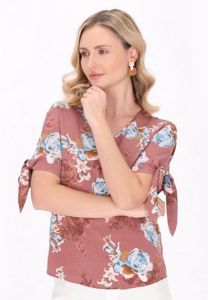 Kobieta o blond włosach w bluzce w kolorze mauve w kwiatowy wzór z marszczonymi rękawami, nosząca okrągłe kolczyki, pozująca z ręką na brodzie patrząc na bok.