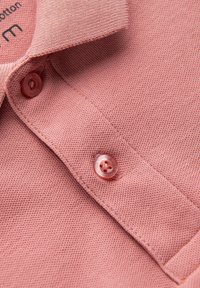 Rosa bomullspolo med en strukturerad väv, med knappslå och runda knappar. Lappen inuti kragen säger "M".