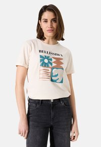  camiseta de algodón color beige claro con un diseño gráfico que presenta patrones en azul, naranja y marrón. Manga corta y escote redondo. Combinada con jeans negros.