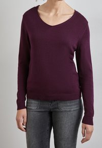 V-ringad tröja i djupt lila tyg, långa ärmar och ribbade muddar och nederkant, kombinerad med mörkgrå jeans. Enkel, figursydd design.