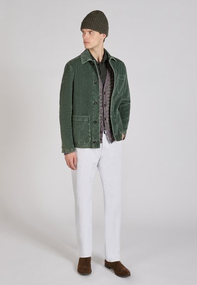 Chaqueta de pana verde con cuello, camisa negra debajo, chaleco estampado, pantalones blancos y botas marrones. Un simple gorro de punto completa el look.