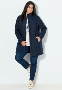 Kvinna i marinblå vadderad jacka, grafisk vit t-shirt, blå jeans och beige sneakers som poserar mot vit bakgrund.