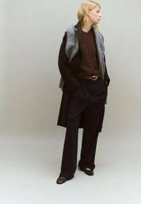 Brauner Zopfmuster-Pullover unter einem schwarzen Mantel, kombiniert mit lockeren schwarzen Hosen und dunkelbraunen Slippern. Grauer Schal über den Mantel drapiert.