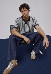 Polo à manches courtes rayé bleu, crème et vert foncé, avec un col en V. Jean décontracté bleu foncé et baskets beiges avec des accents blancs.