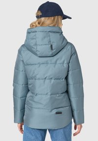 Lichtblauwe pufferjack met een aansluitende capuchon, gestikte textuur en zwart logo accent aan de zoom. Draagt met high-waisted jeans.