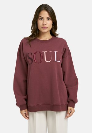 Woman with brown hair wearing a loose maroon sweatshirt with the word "SOUL" in multicolored letters, paired with light beige pants.