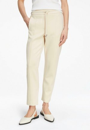 Kobieta w kremowych, dopasowanych spodniach ze ściągaczem w talii i białych, szpiczastych balerinach na pasku, stojąca z rozluźnionymi rękami.