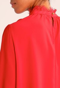 Blusa rossa in tessuto texturizzato, con rifiniture in pizzo sullo scollo e chiusura a bottone sul retro. Le maniche sono svasate.