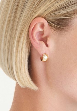Boucle d'oreille en or avec un design incurvé. Surface lisse et brillante ; détail du fermoir visible. Portée dans une oreille percée, affichant un style épuré et moderne.