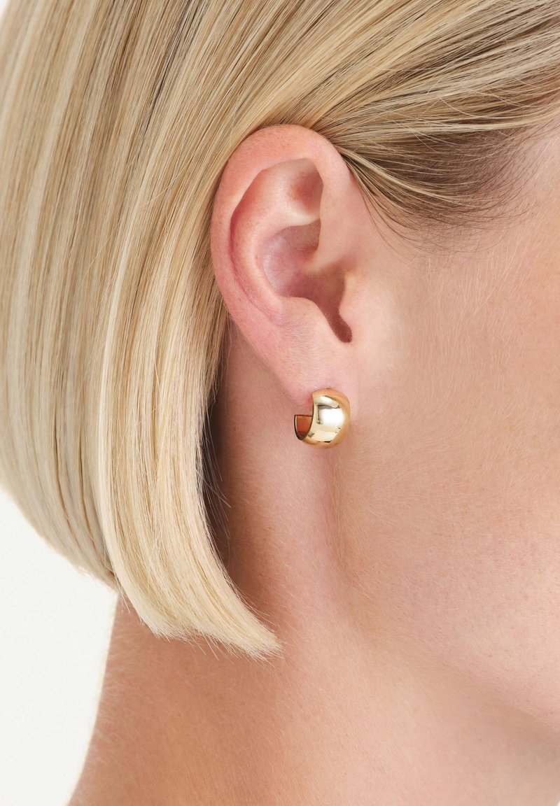 Orecchino a cerchio in oro con design curvo. Superficie liscia e lucida; dettaglio della chiusura visibile. Indossato in un orecchio forato, mostra uno stile elegante e moderno.