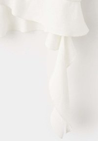 Tissu blanc avec des détails superposés et froncés ainsi qu'une texture douce et fluide suspendu devant un fond clair.