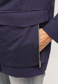 Main dans la poche latérale d'une veste bleu marine avec une fermeture éclair argentée visible, portée sur un pantalon gris clair.