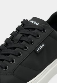 Μαύρα αθλητικά παπούτσια με υφασμένο συνθετικό πάνω μέρος, στρογγυλά κορδόνια, ετικέτα με λογότυπο στη γλώσσα και λευκή καουτσούκ σόλα.