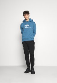 Felpa blu con logo, abbinata a pantaloni jogging neri con testo laterale. Sneakers nere completano l'outfit, il tutto su uno sfondo neutro.