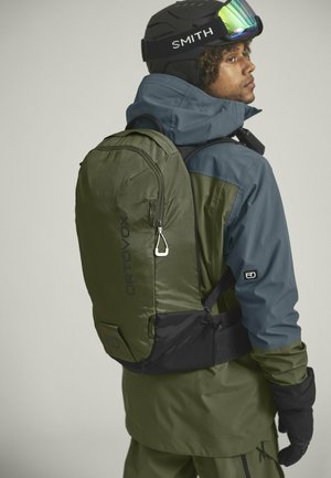 Olivgrüner Rucksack aus Textil, mit einer frontalen Reißverschlusstasche und einem schwarzen Boden. Der Träger trägt eine kontrastierende Jacke in Blau und Grün.
