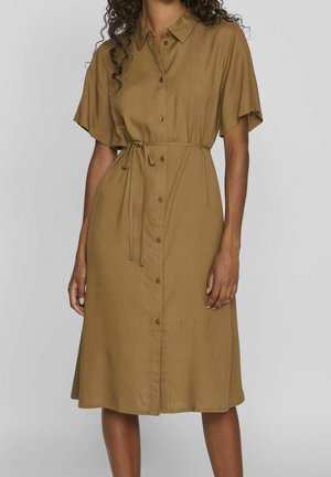 Femme portant une robe-chemise marron aux manches courtes, longueur genoux, avec des boutons sur le devant et une ceinture nouée à la taille.