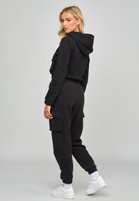 Tuta nera con cappuccio, vita elastica, maniche a polsino e tasche cargo. Abbinata a scarpe da ginnastica bianche, crea una silhouette rilassata.