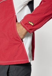 Sarkans un balts vēja aizsargjaka ar elastīgām aprocēm, ar ievērojamu sporta apavu logo uz piedurknes, kā arī sānu kabatām ar rāvējslēdzējiem.