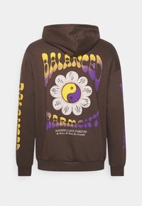 Felpa marrone con il testo "BALANCED HARMONY" giallo e viola, un fiore bianco con simbolo yin-yang e la frase "Niente dura per sempre" sul retro.