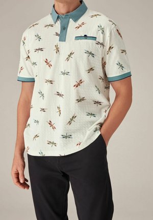 Homme portant un polo blanc à manches courtes avec col et poignets bleus, imprimé de libellules colorées, associé à un pantalon noir.