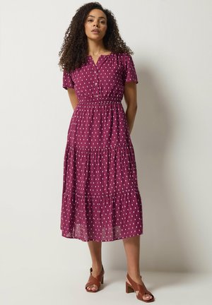 Mujer de pie contra un fondo liso, vistiendo un vestido midi morado de manga corta con botones al frente y un patrón de puntos blancos, junto con sandalias de tacón marrones.