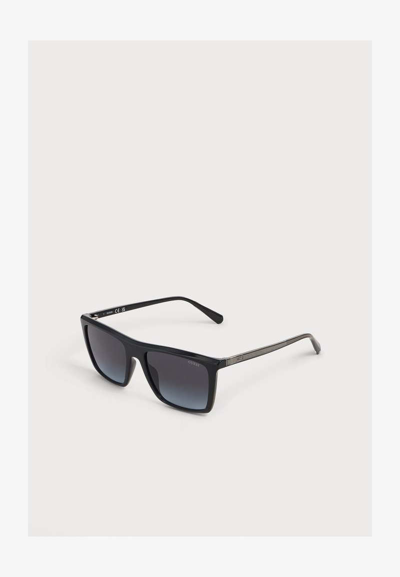 Lunettes de soleil rectangulaires noires avec verres dégradés foncés et branches fines avec un détail métallique près des charnières.
