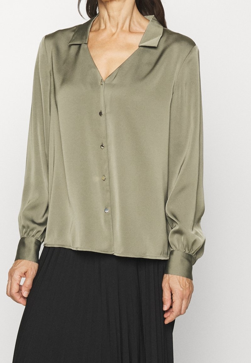 Blouse en satin vert olive avec manches longues bouffantes, col en V et boutons sur le devant. Le tissu présente une texture lisse et un léger éclat.
