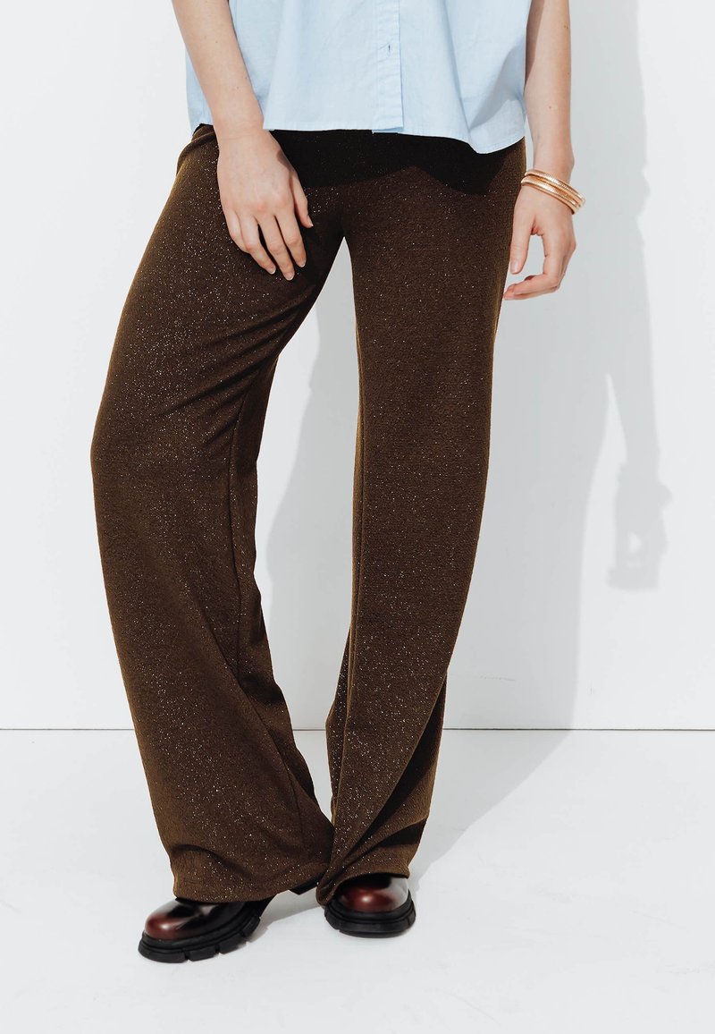 Pantalons bruns évasés en tissu texturé avec des paillettes métalliques, associés à des chaussures à plateforme noires. Une chemise bleu clair est portée au-dessus.