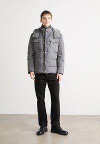Giacca grigia imbottita con colletto alto, due tasche sul petto e bottoni a pressione. Abbinata a pantaloni e scarpe nere, su uno sfondo semplice.