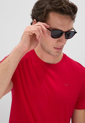 Uomo con maglietta rossa che si sistema degli occhiali da sole neri con la mano destra davanti a uno sfondo chiaro uniforme.