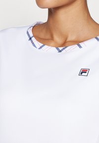 Hvit sportsskjorte med rund halsing og rutete mønster i blått og rødt ved kragen. Fila-logo vises på venstre side.