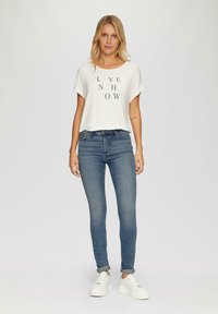 Biely tričko s potlačou "ŽIJ TEĎ", modré skinny jeansy a biele platformové tenisky, stojí na neutrálnom pozadí.