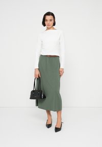 Λευκό μακρυμάνικο τοπ και μακριά φούστα σε ελιά πράσινο, καφέ ζώνη, μαύρη τσάντα χειρός και μαύρα ψηλοτάκουνα παπούτσια με μυτερή μύτη, πάνω σε λευκό απλό φόντο.