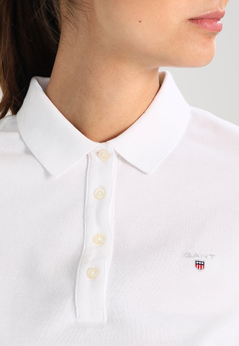 Weißes Poloshirt aus strukturiertem Stoff, mit klassischem Kragen, fünf Knöpfen und einem kleinen gestickten Logo mit roten und blauen Akzenten.