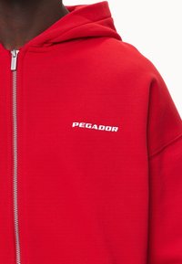 Červená mikina na zip z měkké látky s klokaní kapsou, kapucí se šňůrkami a nápisem "PEGADOR" bíle potištěným na hrudi.