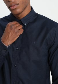 Granatowa koszula na guziki wykonana z bawełny, z klasycznym kołnierzem, długimi rękawami i subtelnym haftem logo na piersi. Tkanina o fakturze.
