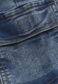 Denimtyg med en blå nyans som har en blekt, sliten struktur. Kontraststygn synliga längs sömmarna och fickorna. Närbild.