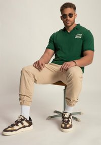 Groene poloshirt met korte mouwen, beige tapered broek en bruine sneakers met witte accenten, zittend op een metalen kruk tegen een lichte achtergrond.