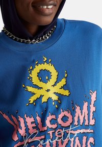 Modrá bavlněná mikina s žlutým plamenem a růžovým nápisem "WELCOME TO" na přední straně, doplněná stříbrným řetízkovým náhrdelníkem.