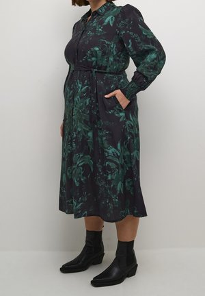 Vrouw draagt een donker midi-jurk met bloemen, lange mouwen, een strik in de taille, zijzakken en zwarte enkellaarzen, staand tegen een effen muur.