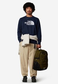 Felpa navy con logo bianco, maglione crema appoggiato, pantaloni cargo beige, scarpe nere e zaino verde oliva con cerniere e tasca in rete.