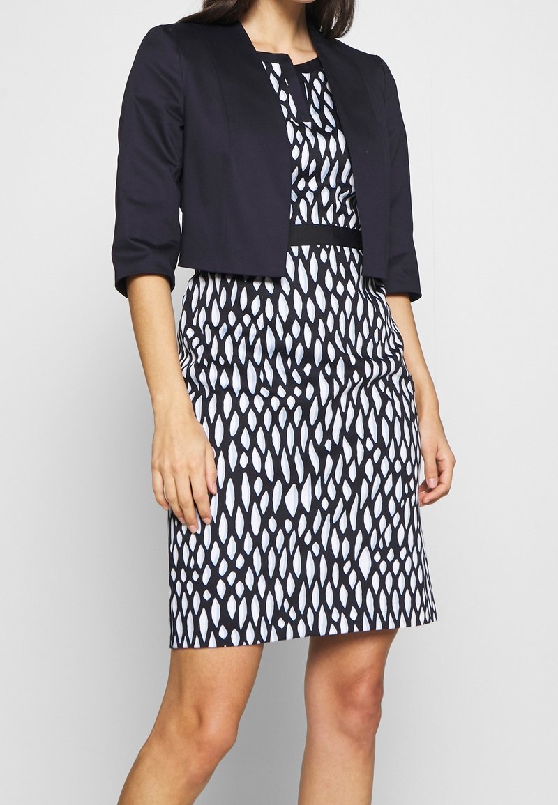 Marineblauwe cropped jas over een zwart-witte jurk met een geometrisch dessin, korte mouwen en een getailleerde silhouet.