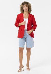 Veste rouge avec une coupe ajustée, un bouton unique sur le devant, deux poches, associée à un short en denim bleu clair et des sandales à talons beiges.