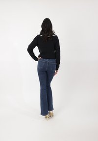 Suéter negro con cuello blanco, combinado con jeans acampanados azules. El conjunto se completa con tacones claros y cabello largo y oscuro.