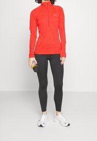 Röd långärmad tröja med fyrdelad dragkedja och tumhål, svarta leggings och vita träningsskor. Håller en vattenflaska.