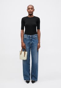 Sort kortærmet top, højtsiddende brede blå jeans, holder en lys mønstret håndtaske med brune håndtag og spidse sorte hæle.
